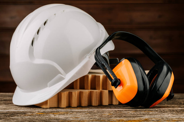 Белый строительный шлем и оранжевые наушники на деревянном фоне. Защитный головной убор
 - Фото, изображение