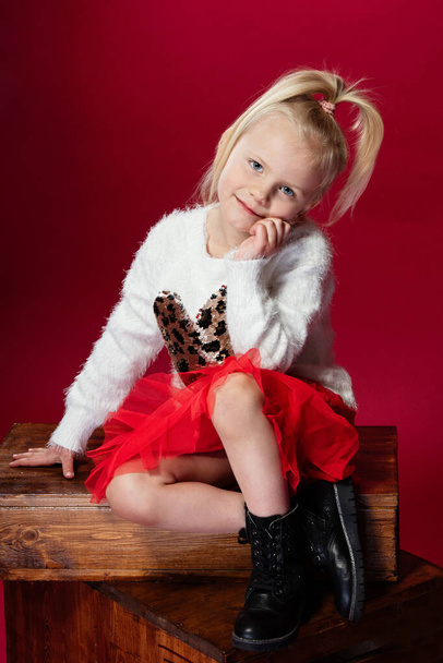 on a red background portrait of a little girl, she sits on boxes of wood and smiles - Foto, Imagen
