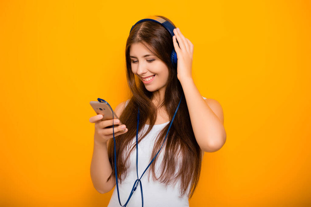 Atraktivní, mladá žena s chytrým telefonem a sluchátky, poslouchá hudbu, zatímco stojí na žlutém pozadí. - Fotografie, Obrázek
