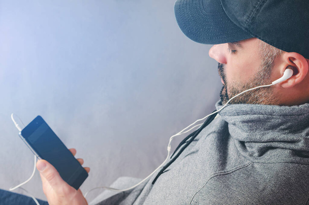 Siyah şapkalı sakallı bir adamın videolar izlediği ya da kulaklıkla akıllı telefondan müzik dinlediği kesilmiş bir portre. Boşluğu kopyala - Fotoğraf, Görsel