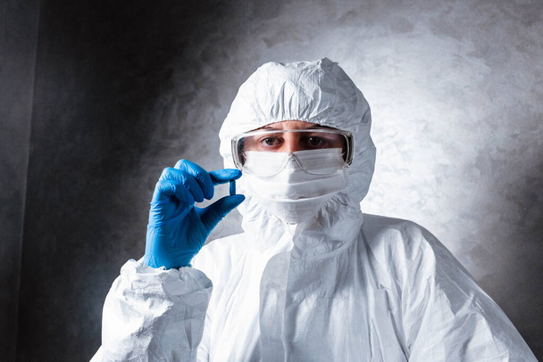 Nurse in a medical face mask and a viral protective suit. The doctor is sitting at the table in a medical face mask and a viral protective suit. The doctor holds a pill in his hands. - Foto, Imagem