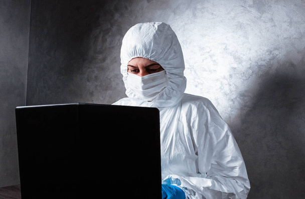 A virologist in a white mask and medical gloves dressed in a white protective suit works with a laptop and looks for information about coronavirus. Isolated and in panic due to epidemic. - Photo, Image