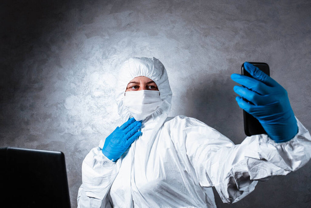 Een jonge vrouw in een viraal beschermend pak met een wit masker en medische handschoenen praat met vrienden tijdens een videogesprek omdat de dokter in quarantaine is geplaatst als gevolg van de verspreiding van het coronavirus epidemie.. - Foto, afbeelding
