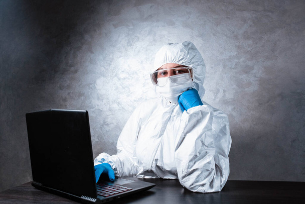 A virologist in a white mask and medical gloves dressed in a white protective suit works with a laptop and looks for information about coronavirus. Isolated and in panic due to epidemic. - Photo, Image