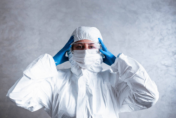 Virologista de máscara branca em um terno protetor viral segura a cabeça, olha para a câmera com medo e pensando em como parar a epidemia de coronavírus e o que fazer. Pensa sobre os perigos disso
. - Foto, Imagem