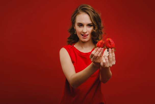 A beautiful girl in a red dress on a red background holds a strawberry in her hands and smiles. - Fotografie, Obrázek