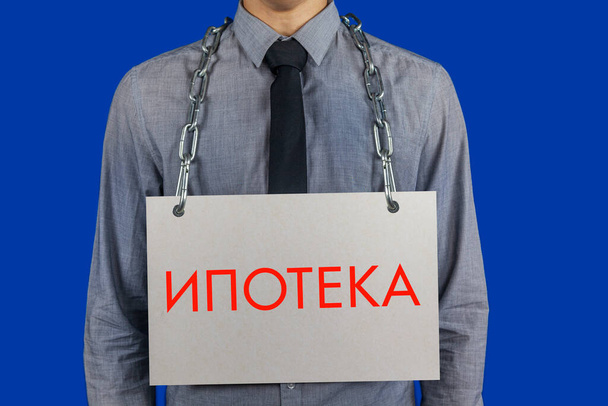 Έννοια: Πίστωση, χρέη, Ένας επιχειρηματίας κρατά μια αλυσίδα γύρω από το λαιμό του με μια αφίσα με την επιγραφή στο πιάτο στη ρωσική "υποθήκη" - Φωτογραφία, εικόνα