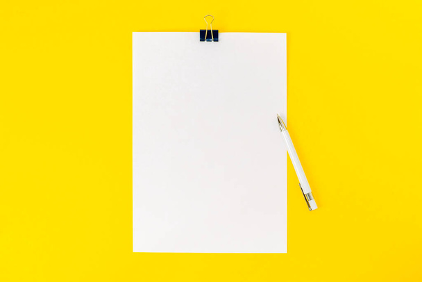 Een leeg vel papier is bevestigd met een paperclip en een witte pen op een centrum van gele achtergrond. Mockup, blanco voor aankondiging, informatie, verklaring, school. Bovenaanzicht, vlak lay. - Foto, afbeelding