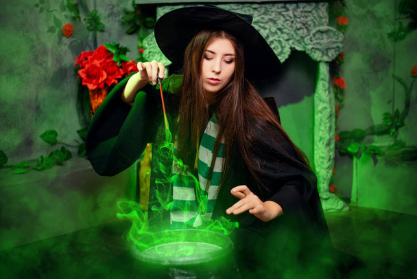 A young witch in a black hat and mantle makes a potion in a black cauldron with a magic wand and a book. A girl sits on the floor in magical green smoke. - Foto, Bild