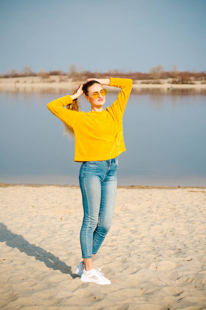 Блондинка в желтом свитере, синих джинсах, на пляже на фоне голубого неба и воды в очках, прикасаясь к волосам
 - Фото, изображение