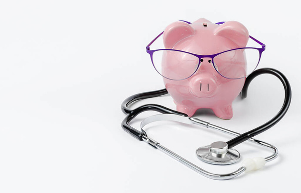 白い背景に眼鏡をかけた聴診器と貯金箱。医療保険適用の概念 - 写真・画像