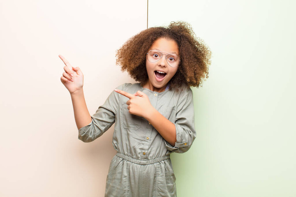 african american little girl feeling joyful and surprised, smiling with a shocked expression and pointing to the side against flat wall - Fotó, kép