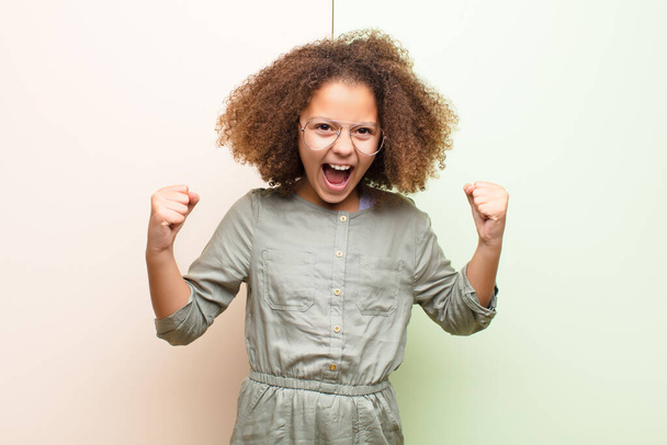african american little girl feeling happy, positive and successful, celebrating victory, achievements or good luck against flat wall - Фото, зображення