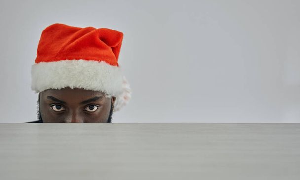 De Afrikaanse man in de kerstmuts die toekijkt vanaf de tafel. - Foto, afbeelding