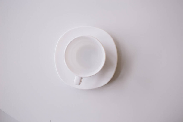 Ένα λευκό φλιτζάνι για τσάι ή καφέ στο λευκό πιάτο στο λευκό τραπέζι ή φόντο. επίπεδη θέσει. νεκρή φύση - Φωτογραφία, εικόνα