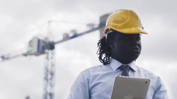 Πορτρέτο ενός Αφρικανού με καπέλο χρησιμοποιώντας tablet στο εργοτάξιο και παρατηρώντας το έργο - Πλάνα, βίντεο