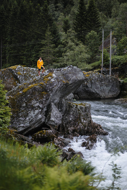 A guy in a yellow raincoat standing on a rock against the river - Photo, image