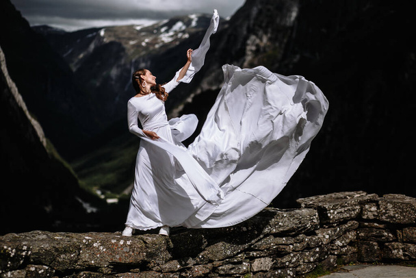 A bride standing on the edge of a stone wall and waving the hem of her wedding dress - Fotografie, Obrázek