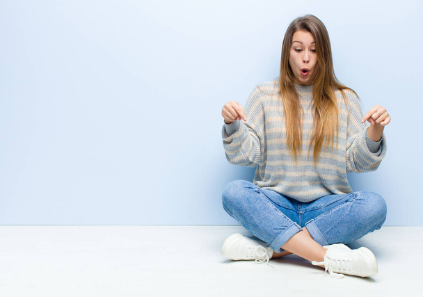 young blonde woman feeling shocked, open-mouthed and amazed, looking and pointing downwards in disbelief and surprise sitting on the floor - Photo, image