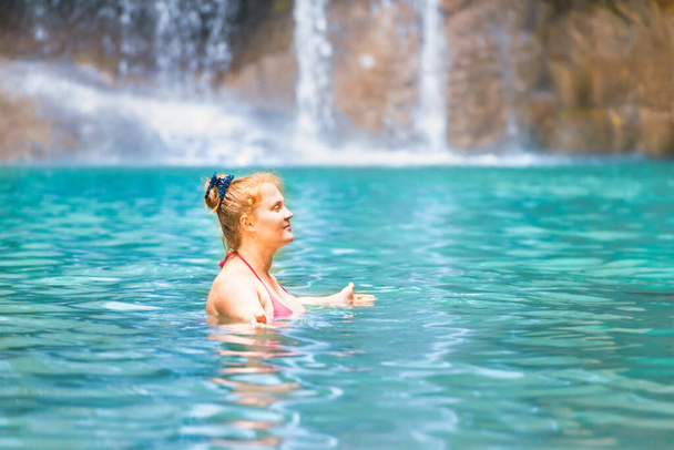 Red haired young woman in pink bikini swimsuit relaxes in emerald tropical lake with waterfall. Erawan National park, Kanchanaburi, Thailand - 写真・画像