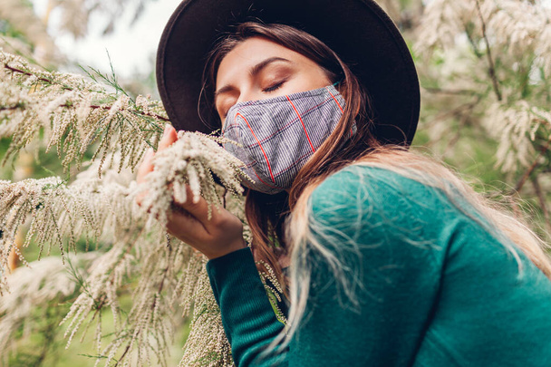 La chica huele flores en el parque. La mujer lleva máscara en el jardín durante la pandemia del coronavirus covid-19. Moda verano look
 - Foto, Imagen