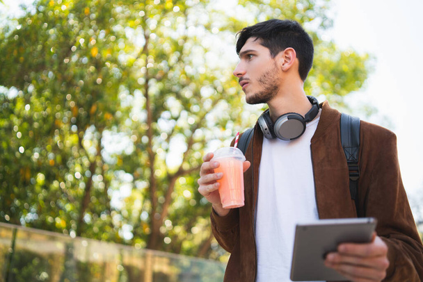 Portrait of young man using his digital tablet while drinking fresh fruit juice outdoors in the street. Technology and urban concept. - Foto, Bild
