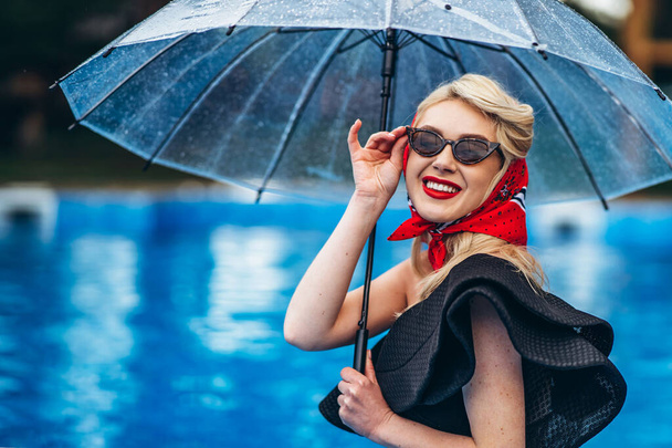 Pin up styled blonde in sunglasses, with red lips and reto hairstyle, in black vintage swimsuite and red scarf, holding umbrella and have fun in the swimming pool - Foto, Imagen