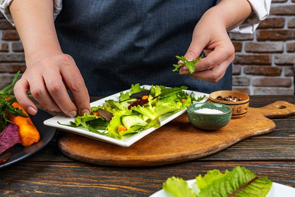Womans hands decorate and prepare a fresh green salad in a plate with spinach, arugula, carrot romance, on an old wooden table. Natural healthy food concept. - Photo, image