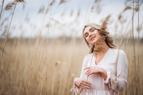 Photo of a pretty smiling girl with long blond curly hair in light long drees standing in a reed field and holding high reed branches in her hands - Fotografie, Obrázek