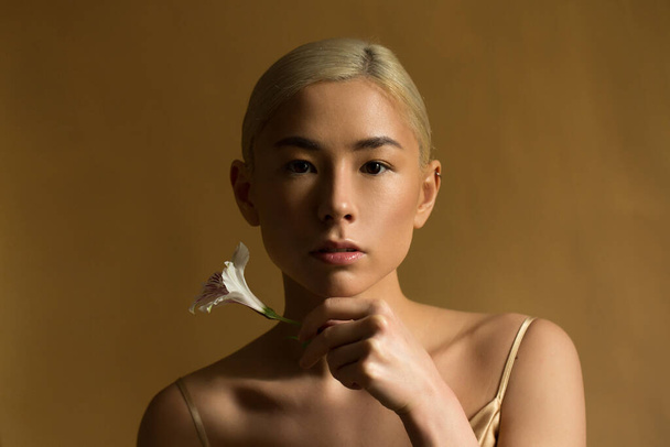 Beautiful woman on a dark yellow background looks straight and holds a flower in her hands. Low key. Blond hair, brown eyes. Brush near the chin. - Foto, imagen
