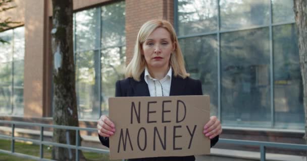 Frustrated business woman in 30s getting fired and showing carton banner with need money writing. Female worker lost job and standing at street. Concept of financial crisis and unemployment. - Filmagem, Vídeo