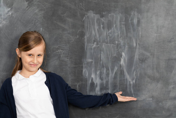A girl stands smiling at a chalkboard and points to an empty space ready for writing with chalk. - Foto, immagini