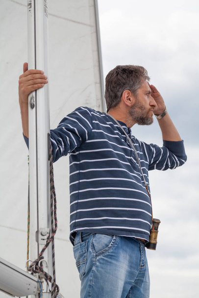 captain holds on to the mast. A man in a striped sweater and jeans sits on the nose of a sailing yacht and looks through binocular - Foto, immagini
