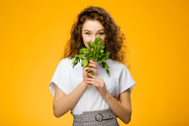 Closeup portrait of pretty caucasian girl with curly hair holding green fresh parsley isolated on colorful yellow background - Фото, изображение