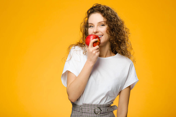 Closeup portrait of pretty caucasian girl with curly hair and red apple isolated on colorful yellow background - Valokuva, kuva