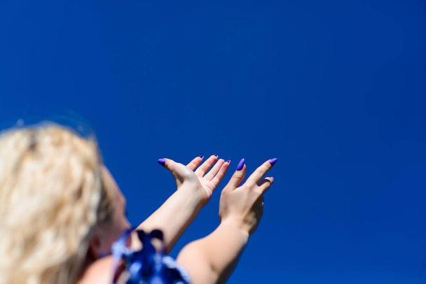 A young blonde girl is looking at the beautiful blue sky and reaches for the sky with her hands. Beautiful female admires nature. - Foto, Bild