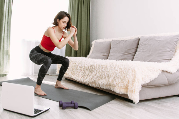 Mujer hermosa joven hace sentadillas y sacude los músculos glúteos en casa, repitiendo entrenamiento en línea. Deportes de cuarentena
. - Foto, imagen