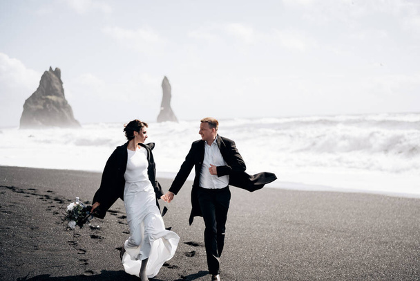 Προορισμός Ισλανδία γάμος. Το ζευγάρι των γάμων τρέχει κατά μήκος της αμμώδους μαύρης παραλίας του Vik, κοντά στο βράχο βασάλτη, με τη μορφή πυλώνων. Κρατώντας τα χέρια, τρέχοντας κατά μήκος της ακτής με φόντο τα κύματα - Φωτογραφία, εικόνα