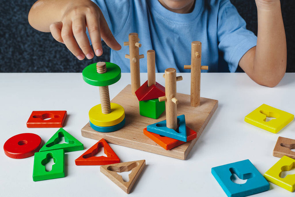 Child plays Montessori game. Kid collects wooden toy sorter. Multicolored geometric shapes, circle, square, triangle, rectangle. - Фото, изображение