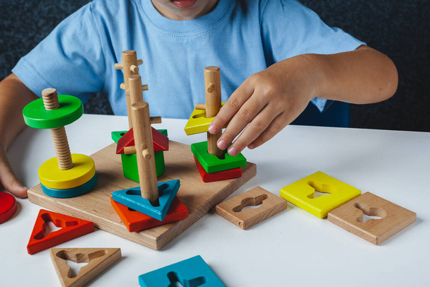 Child plays Montessori game. Kid collects wooden toy sorter. Multicolored geometric shapes, circle, square, triangle, rectangle. - 写真・画像