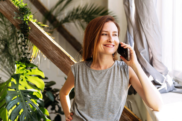 Een mooie vrouw spreekt thuis aan de telefoon met familie of vrienden op een achtergrond van diverse planten. - Foto, afbeelding