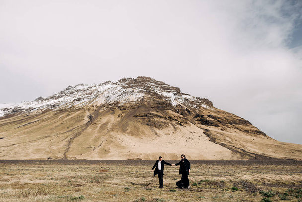 Προορισμός Ισλανδία γάμος. Γαμήλιο ζευγάρι σε φόντο χιονισμένων βουνών. Η νύφη και ο γαμπρός με τα μαύρα παλτά αγκαλιάζονται σε ένα χωράφι με βρύα και κίτρινο γρασίδι.. - Φωτογραφία, εικόνα
