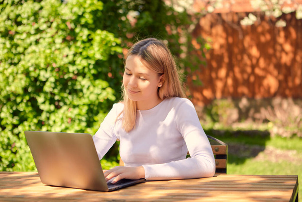 A cute young teen girl is learning online on a laptop while sitting at a wooden table in the back garden of a house. - Foto, immagini