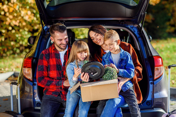 Lovely young parents with their cute kids sitting in the trunk and holding carton box with plants and other home things during moving into new apartment. - Foto, afbeelding