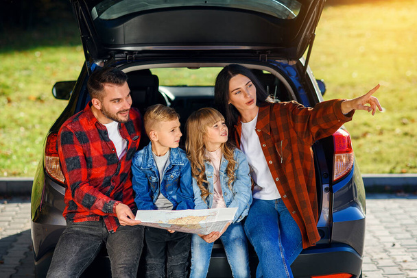 Satisfied family of handsome father, pretty mother and lovely kids which are looking on the road map while sitting in the cars trunk at joint family vacation. - 写真・画像