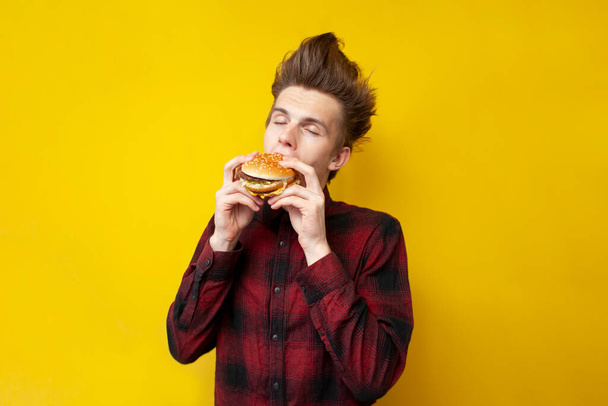 young shocked guy eating a burger on a yellow isolated background, hipster with funny hairstyle holds fast food - Photo, Image