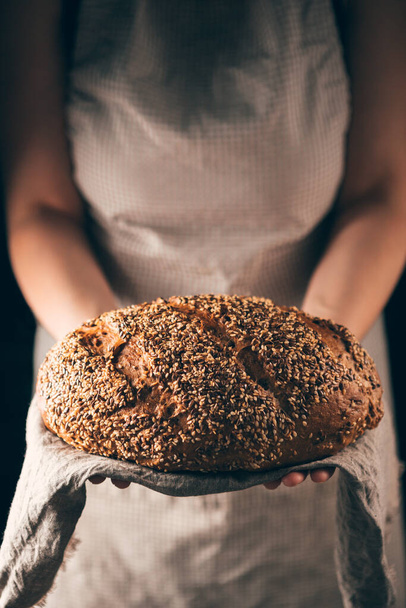Female hands holding loaf of homemade rye bread with sesame and linseeds on towel. Concept for bakery poster or cover - Fotó, kép