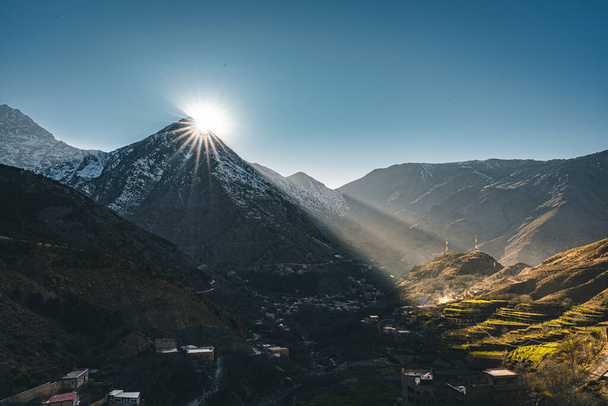 Widok zachodu słońca z gwiazdą słońca w wiejskiej górskiej wiosce Imlil w wysokich górach Atlas Maroko w Afryce. Pokryte śniegiem szczyty w tle z latarniami i błękitnym niebem. - Zdjęcie, obraz