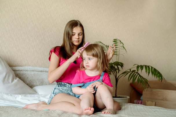Lapset leikkivät sängyllä valoisassa makuuhuoneessa. Teinityttö kampaamassa vauvan hiuksia. Hauskat tytöt. Sisarellinen rakkaus. loma-asunto - Valokuva, kuva