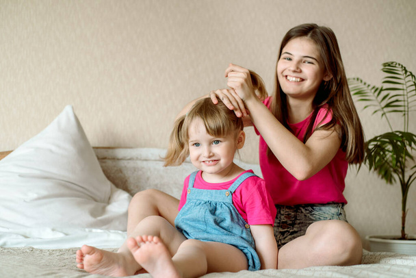 Children play on the bed in a bright bedroom. teen girl combing baby hair. funny girls. sisterly love. vacation home - Photo, image